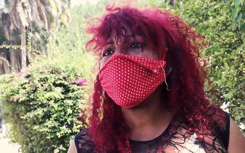 A Venezuelan woman with bright red hair and a red mask stands at the edge of a road.