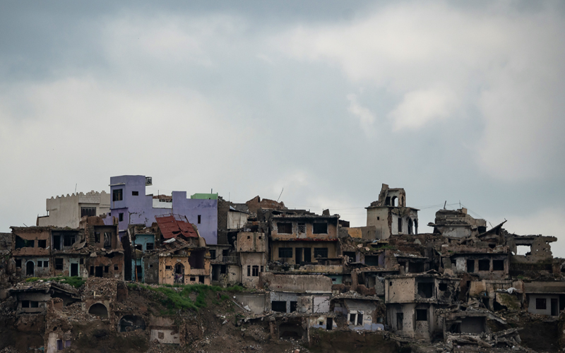 A series of buildings in an urban area with roofs caved in and bullet-ridden walls crumbling, some sinking into the ground.