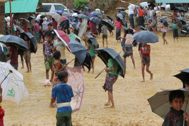 Several people walk barefoot on a flooded terrain in a Rohingya refugee camp.