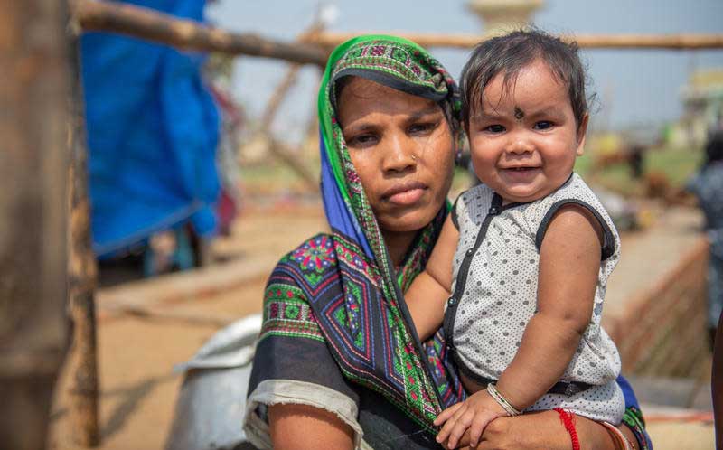 In India, a woman in a green and blue sari holds her young child who smiles at the camera