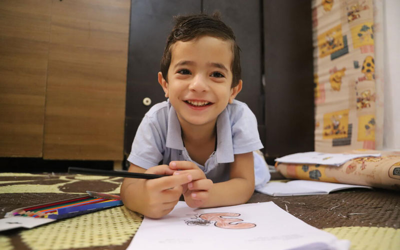 A boy smiles while lying on the floor, colouring illustrations on paper.