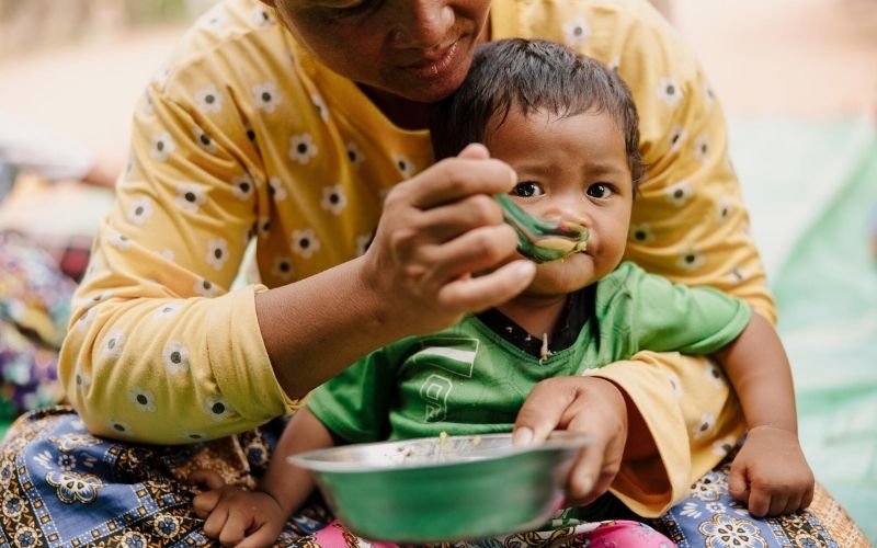 A toddler sits in her mother's lap as she is being fed from a metal bowl.