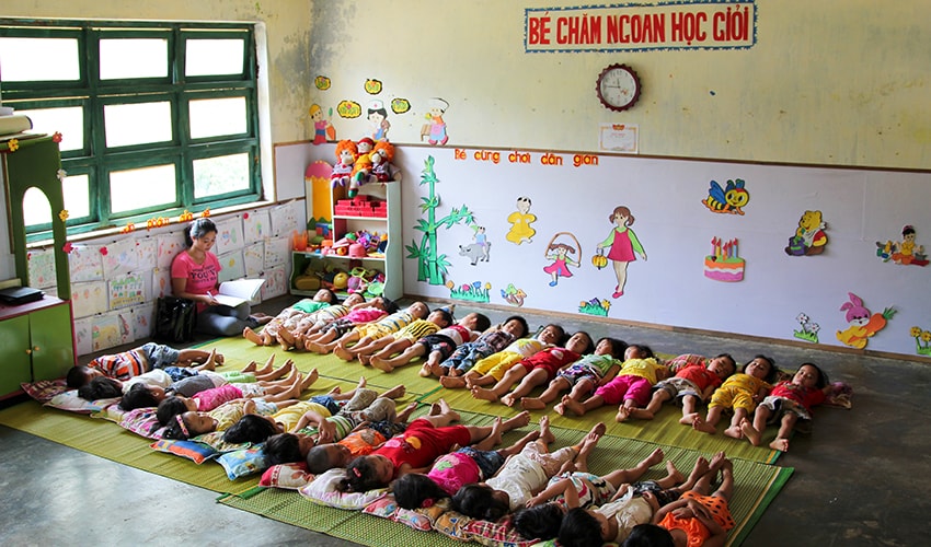 In a classroom in Vietnam, toddlers lie down side by side for naps, while their teacher reads close by.