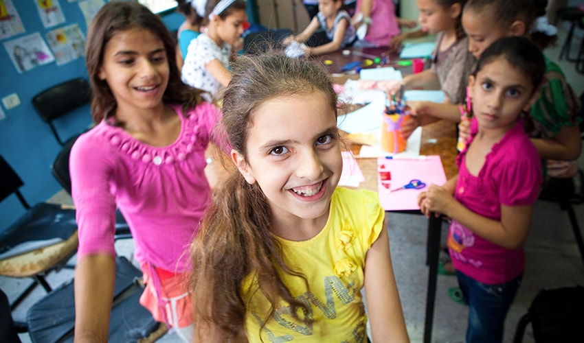 Three girls sit together with other students and work on crafts in the West Bank. One girls smiles at camera.