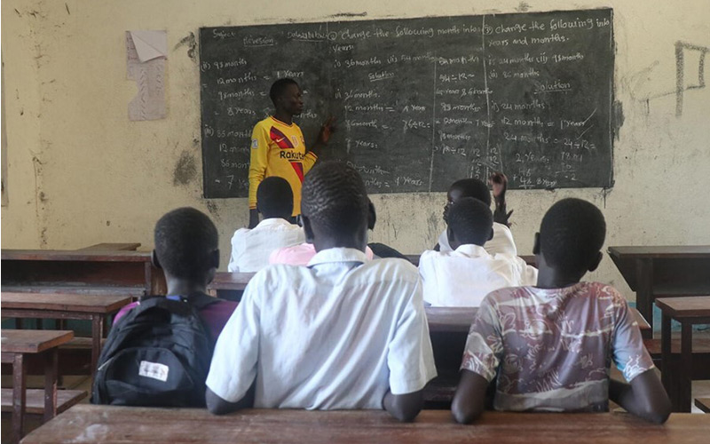 A teacher stands in front of a group of boys inside a classroom.