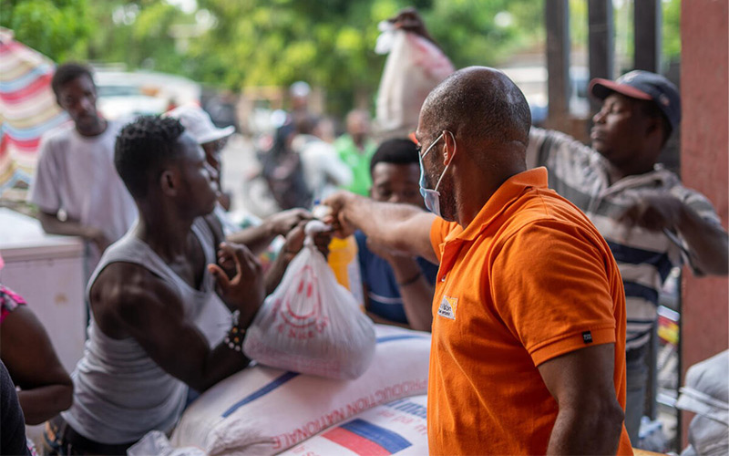 A man in orange shirt hands out a bag of food. items to another man.