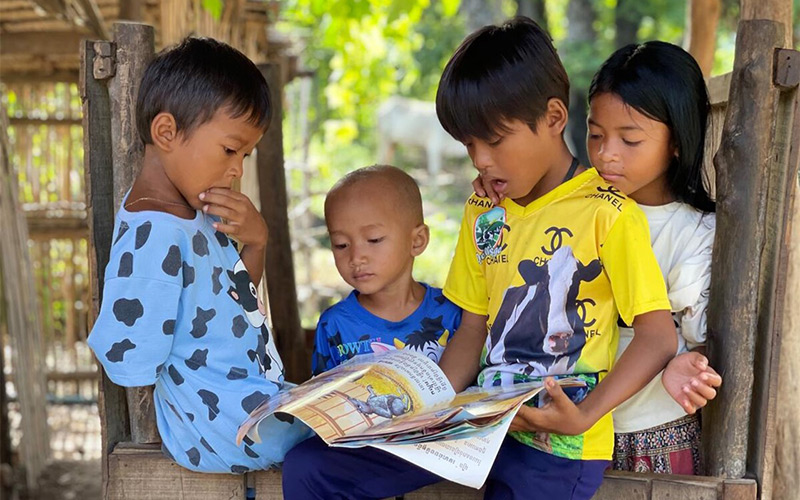 a boy reads a book while three other children stand around him, listening.