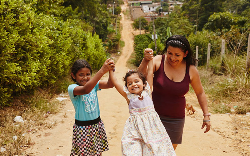 a woman and two children laugh and hold hands walking down a dirt road.