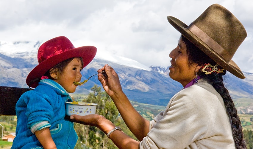 A Peruvian mother feeds her toddler daughter from a spoon. They on on a mountain top and the Andean mountains can be seen in the background.