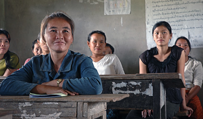 In a classroom in Laos, mothers sit at desks and look toward the front of the room.