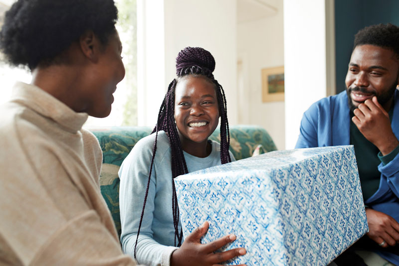 Three young adults sitting on a sofa, while one happily holds on to a large wrapped present.