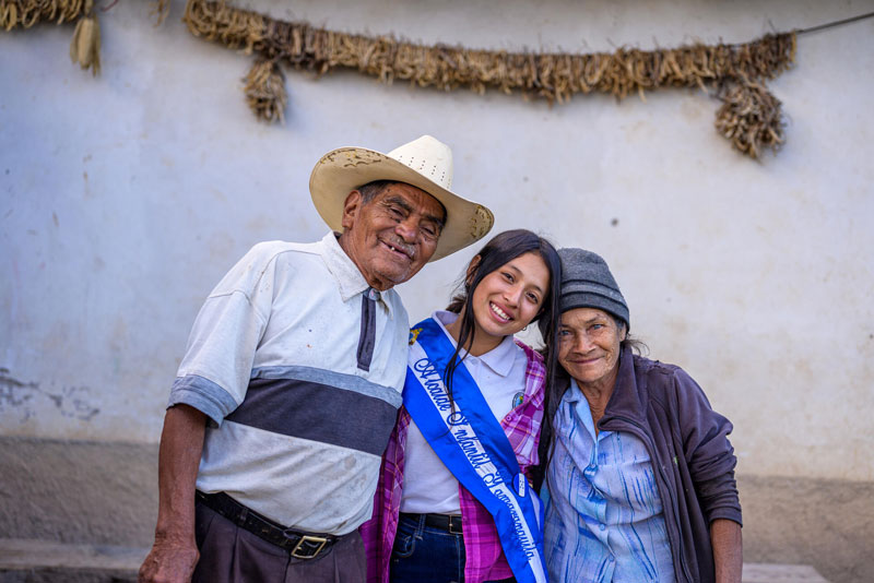 A teenage girl wearing a blue-ribbon sash stands in front of a concrete wall with two elderly people as they embrace and smile for a picture.