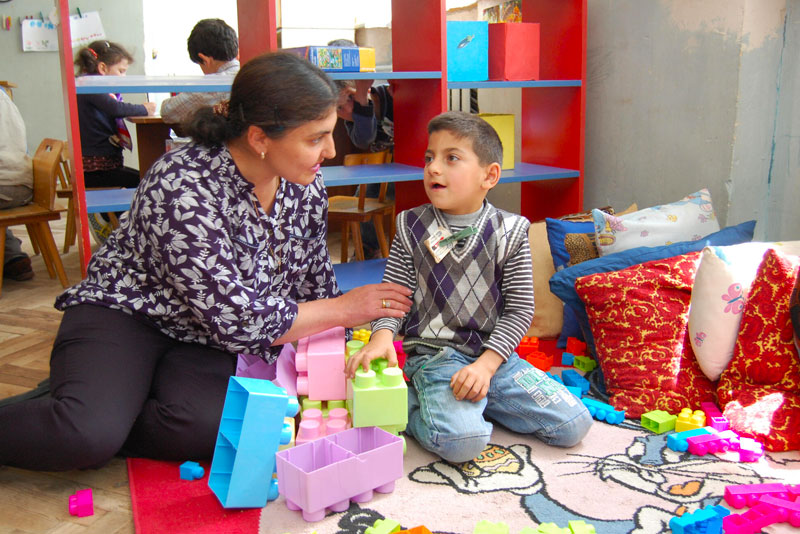 An adult woman is seated on the ground beside a young boy, assisting him as he plays with toys in the learning center.