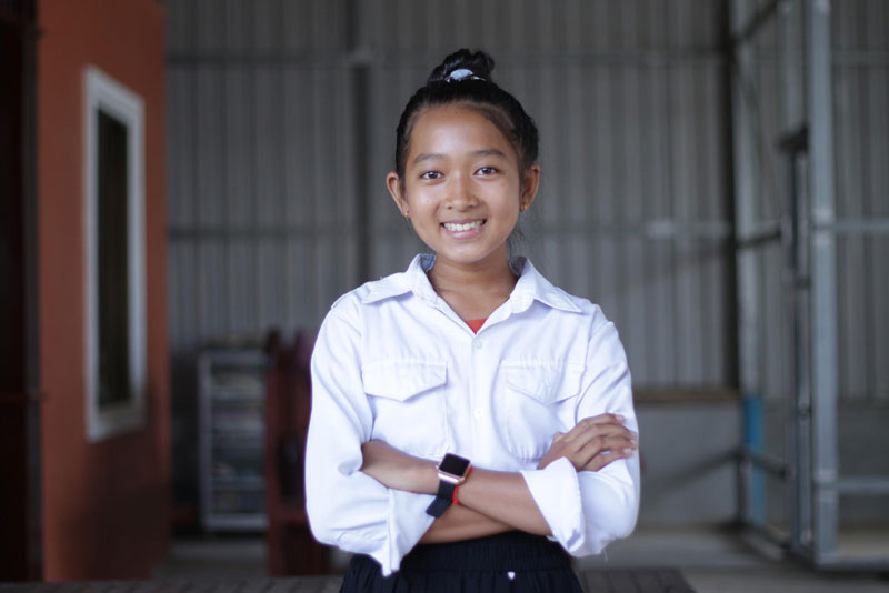 A young girl in a white collared shirt stands with her arms crossed, smiling for a picture.