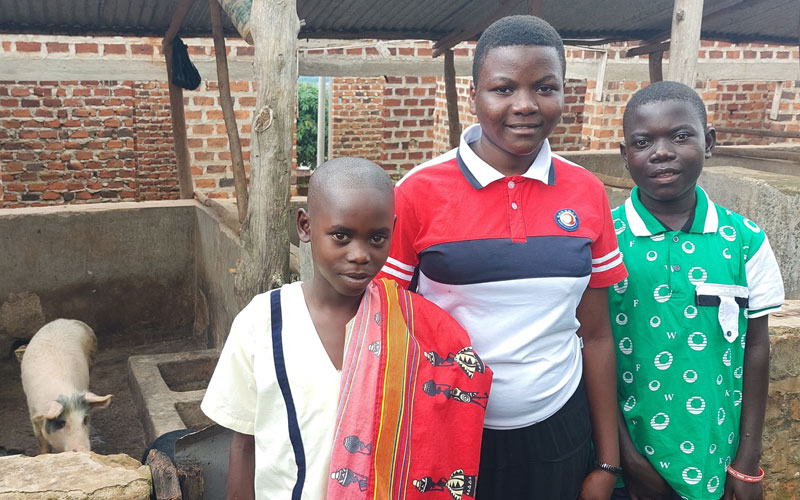 Three sponsored children from Uganda stand beside one another in front of a pig pen.