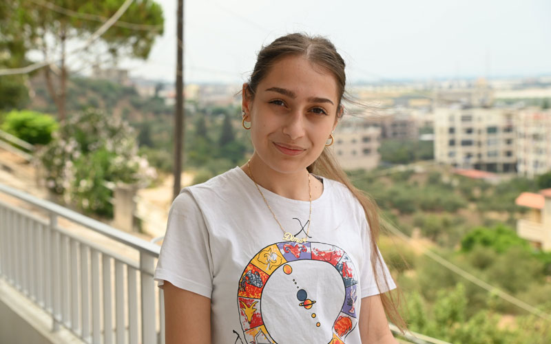 A young woman wearing a white t-shirt and gold necklace stands on a balcony and smiles softly at the camera.