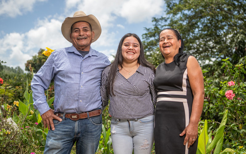 A father, young woman, and mother stand together as they smile in front of a garden.