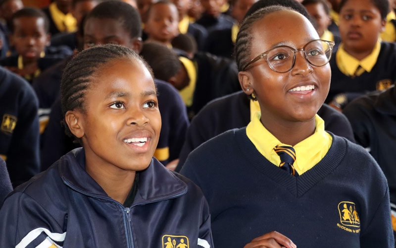 Two school girls in uniforms are seated at a school-wide assembly. They are smiling as they listen attentively.