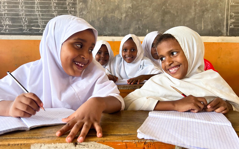 Young school girls dressed in Hijab’s smile at each other as they work at their desks.