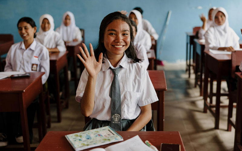 A student seated in a classroom desk smiles as she raises her hand.