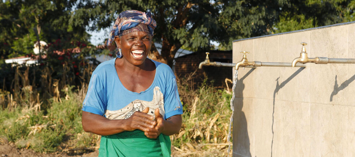 A woman smiles joyfully as she collects water from an outdoor tap source.