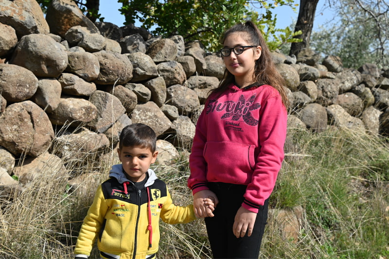 A girl and her little brother hold hands and stand in front of a stone wall, smiling at the camera.