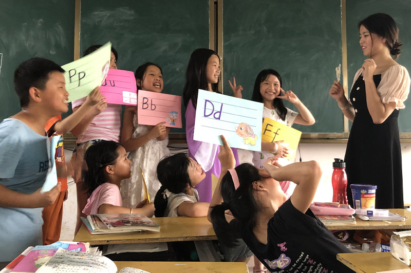 Standing on the right, Fang faces her students who are holding up colourful pages with the alphabet written on them.