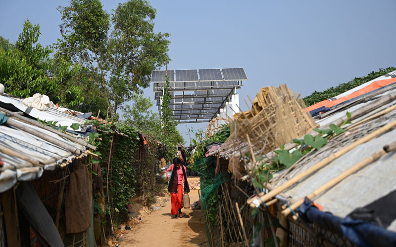 a young Rohingya girl carries water down a dirt path lined with crude shelters with tall solar panels behind her