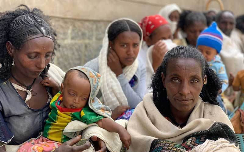 A long line of women, several holding infants, sit against a wall at a food distribution centre.