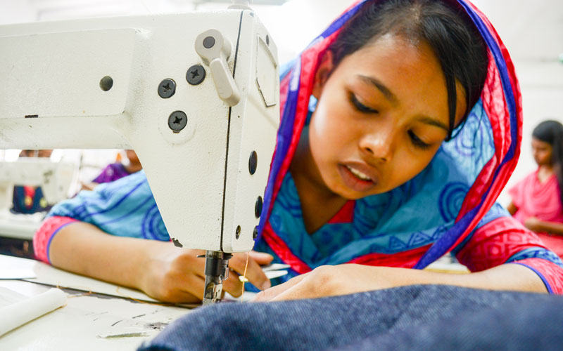 A young girl leans her face close to a sewing machine needle as she stitches denim jeans.