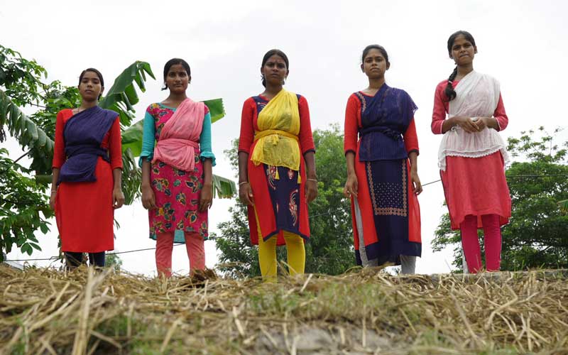Five young Indian women stand in a line on a road, wearing sombre expressions.