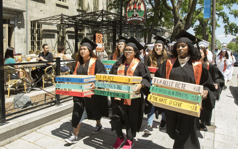 A group of students march down a sidewalk wearing black graduation caps and gowns.