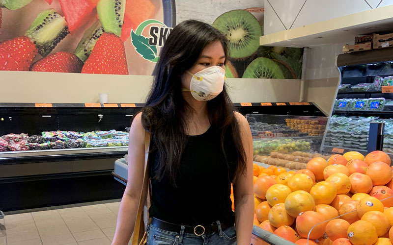 A young Asian-Canadian woman shops for oranges at the grocery store. She is wearing a mask.
