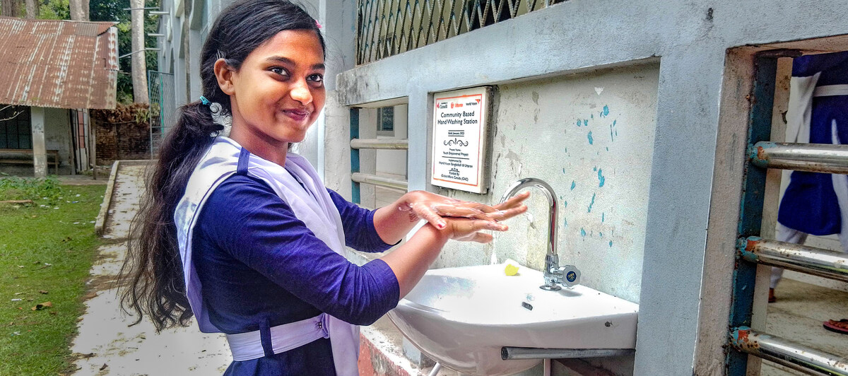 A young girl smiles for the camera as she washes her hands at a wash station.