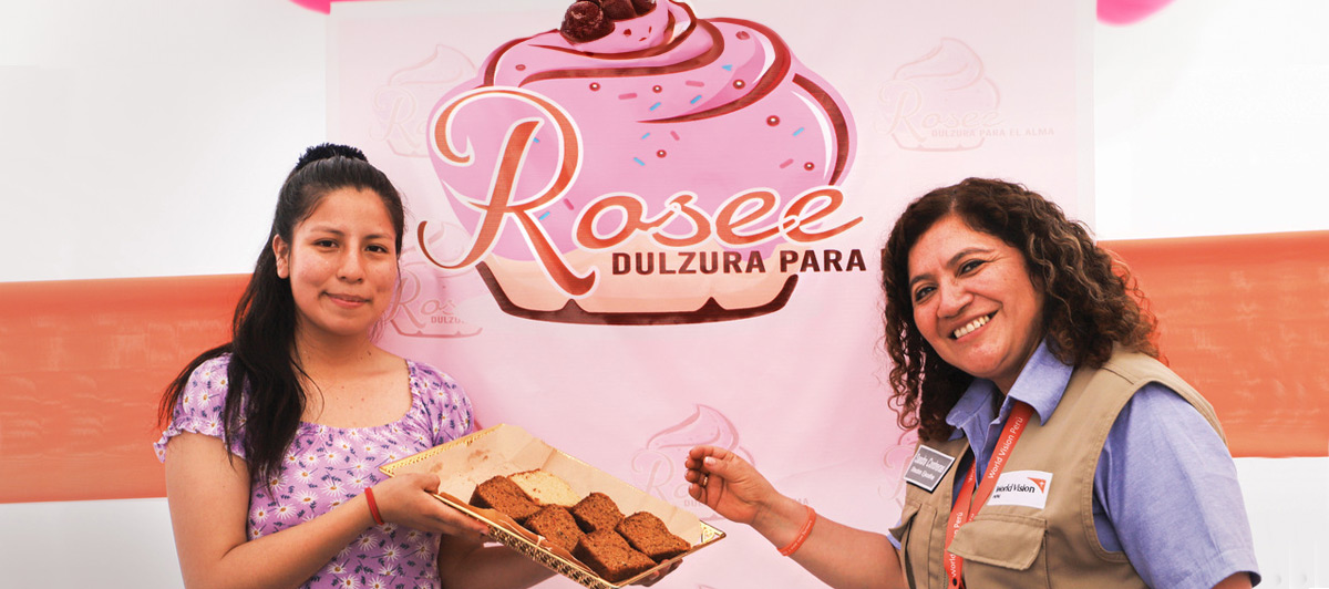 Two women smile as they stand in front of a colourful commercial sign. The woman on the left holds a tray of baked goods.