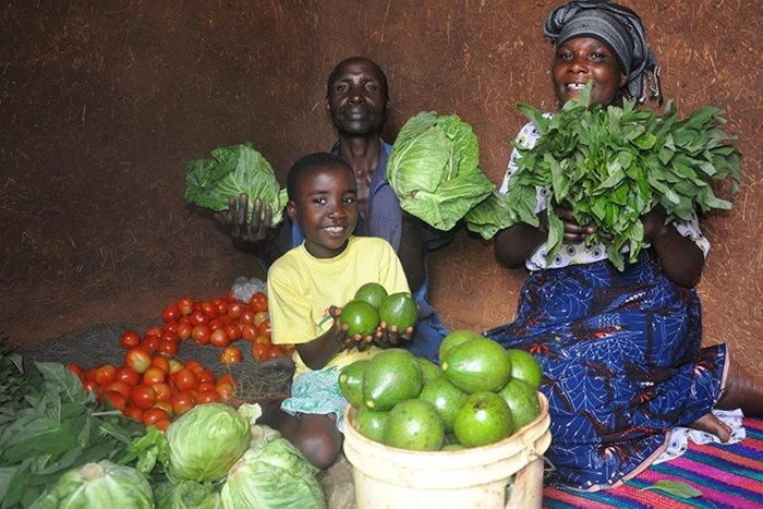 In Shishiyu, Tanzania, a man, woman and young boy sit on a carpet holding up various vegetables.