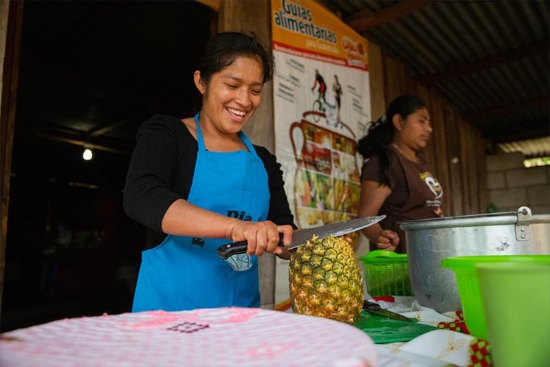 In San Pedro, Guatemala, a woman in a blue apron chops a pineapple as another woman stands by.