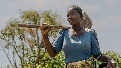 A woman in a blue shirt carrying a hoe and watering can outdoors, surrounded by greenery.