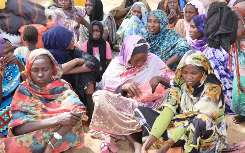 A group of people sit closely together outdoors, wearing colorful patterned clothing and head coverings.