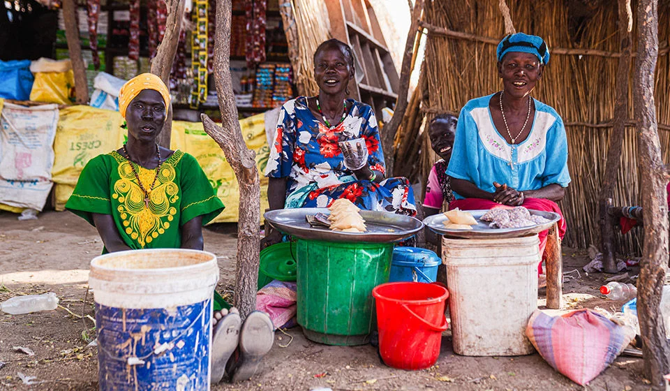 Three women sit surrounded by colorful plastic containers at a market.