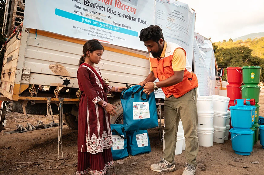 World Vision staff handing hygiene kit to a girl beside a truck displaying disaster‑response banners in a rural area.