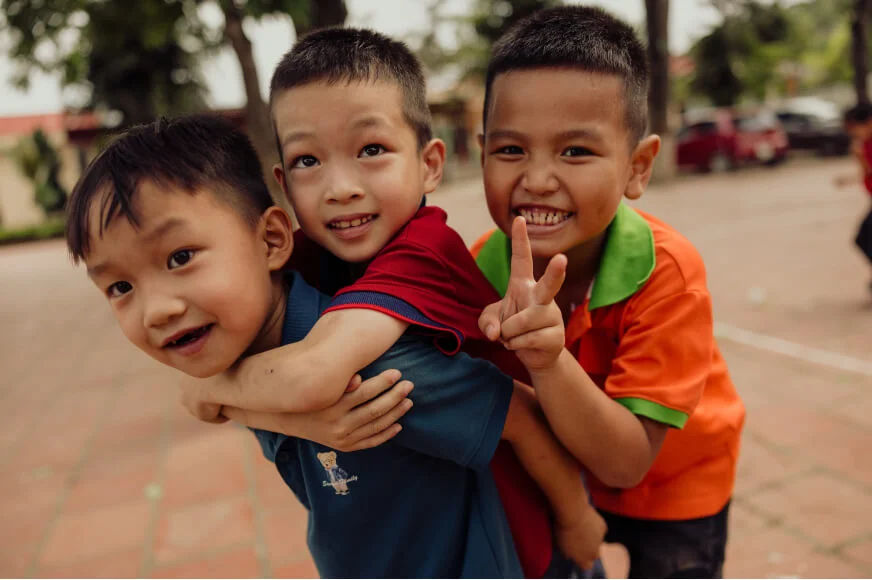 A little boy wearing a red shirt clings to an older boy’s back as another boy in an orange shirt makes a peace sign behind them.