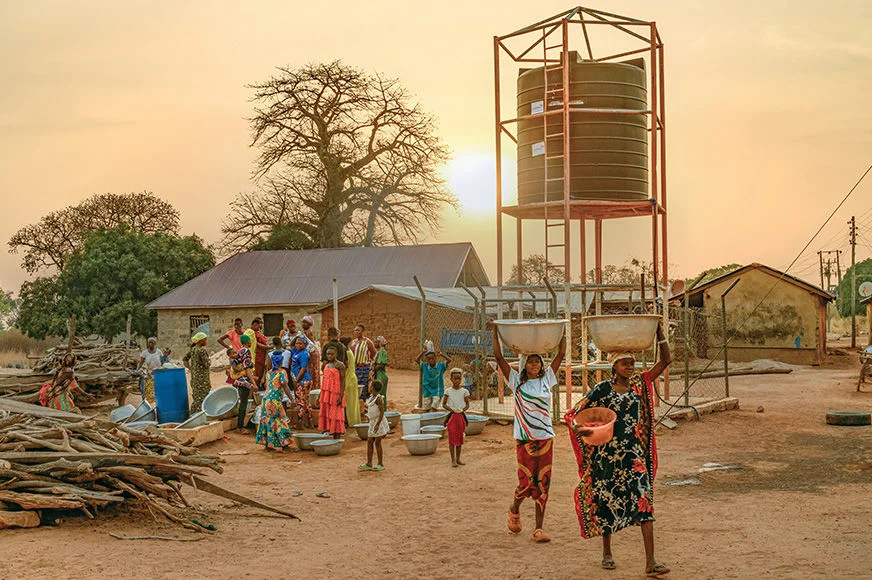 An enormous water tank towers over a group of houses as women, men and children gather around to fill large metal bowls and buckets with clean water. (Ghana, 2025)