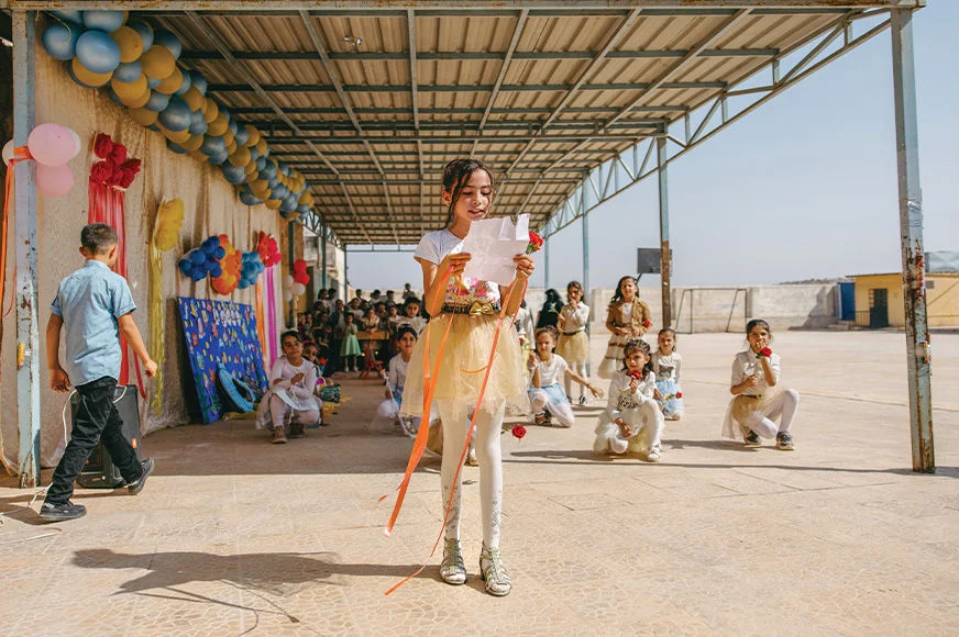 A young girl wearing a white shirt and yellow skirt is reading a letter after participating in a community event. (Syria, 2024)