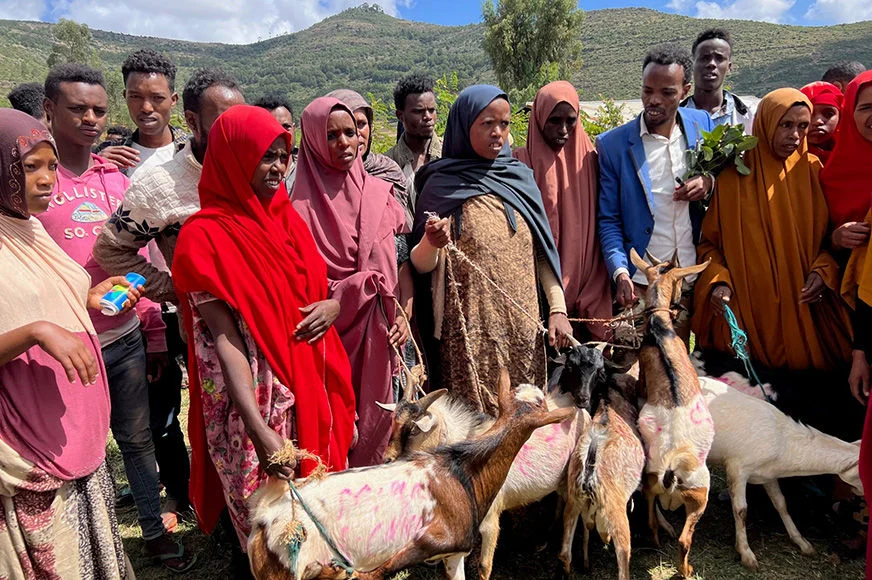 A group of people gather with goats in a rural area, celebrating or participating in an event.
