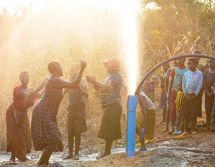 A group of people dancing and celebrating as water sprays into the air from a well.