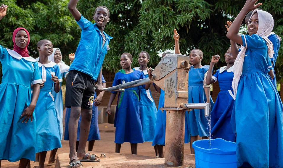 School students in blue and white uniform dancing around a water pump that is filling up a bucket. (2025)