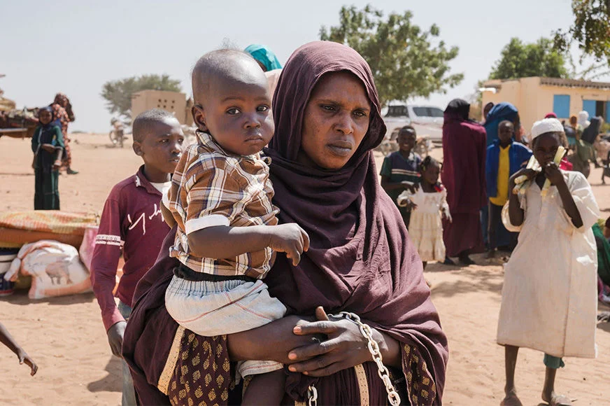 A mother holds a young child while standing among other families at the Sudan-Chad border.