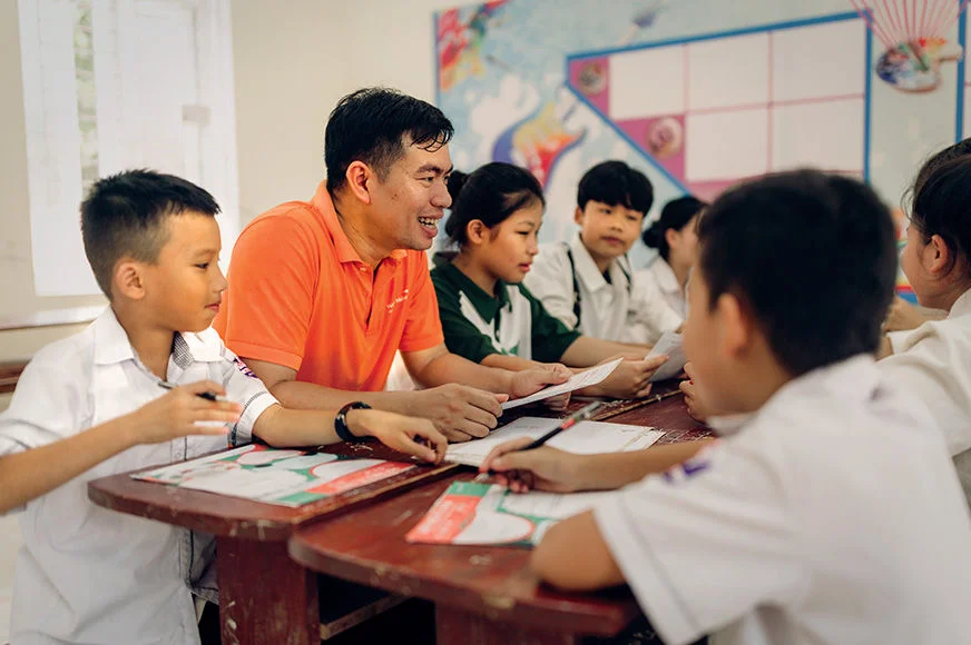 A male teacher wearing an orange t-shirt is sitting with his young students as they participate in a classroom activity. (Vietnam, 2024)
