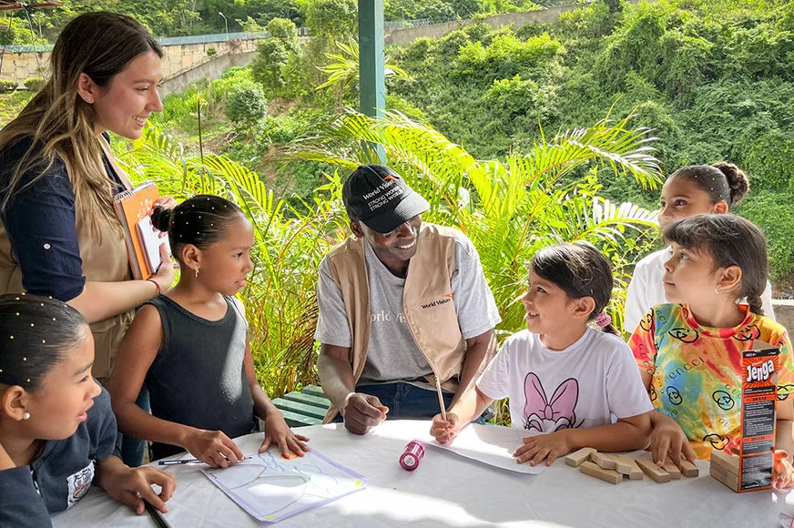 World Vision staff leading outdoor educational activity with children, featuring drawing materials and games on a table in a lush, green setting. 